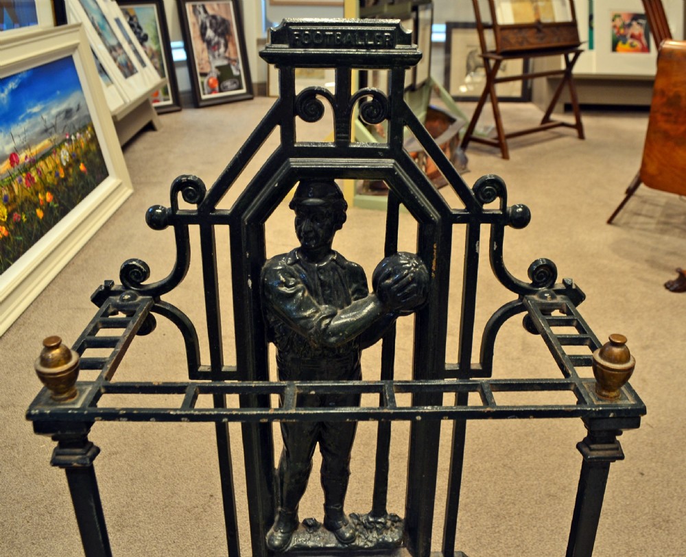 A black cast iron stick stand featuring a central relief figure of a nineteenth-century footballer holding a ball, framed by scrolling detail and twin drip trays. View V