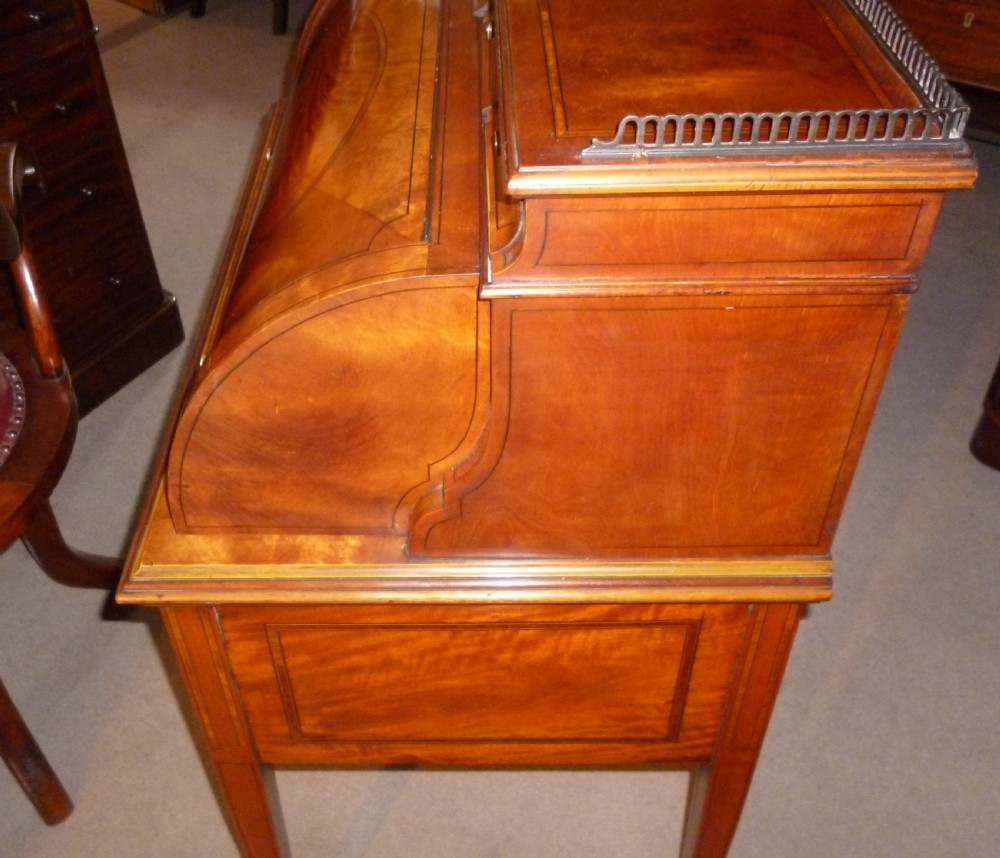 An Edwardian satinwood rolltop bureau featuring a cylinder front with an oval marquetry panel, a brass gallery rail top, and tapered legs. View IV