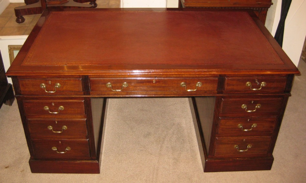 An antique mahogany pedestal desk featuring nine drawers with brass handles and a reddish-brown leather writing surface.