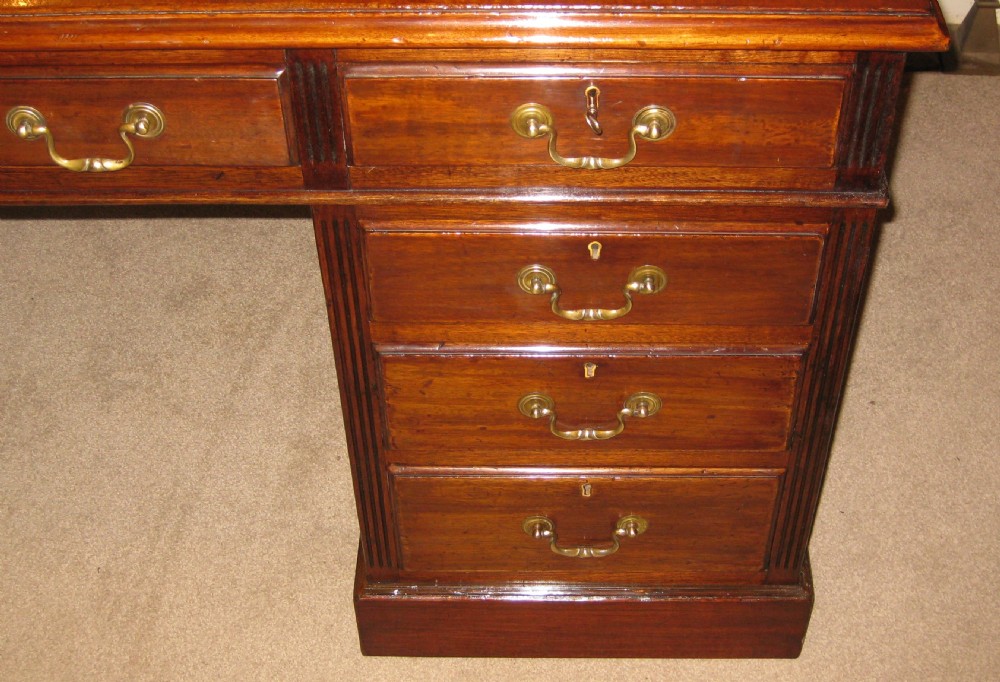 An antique mahogany pedestal desk featuring nine drawers with brass handles and a reddish-brown leather writing surface. View VI