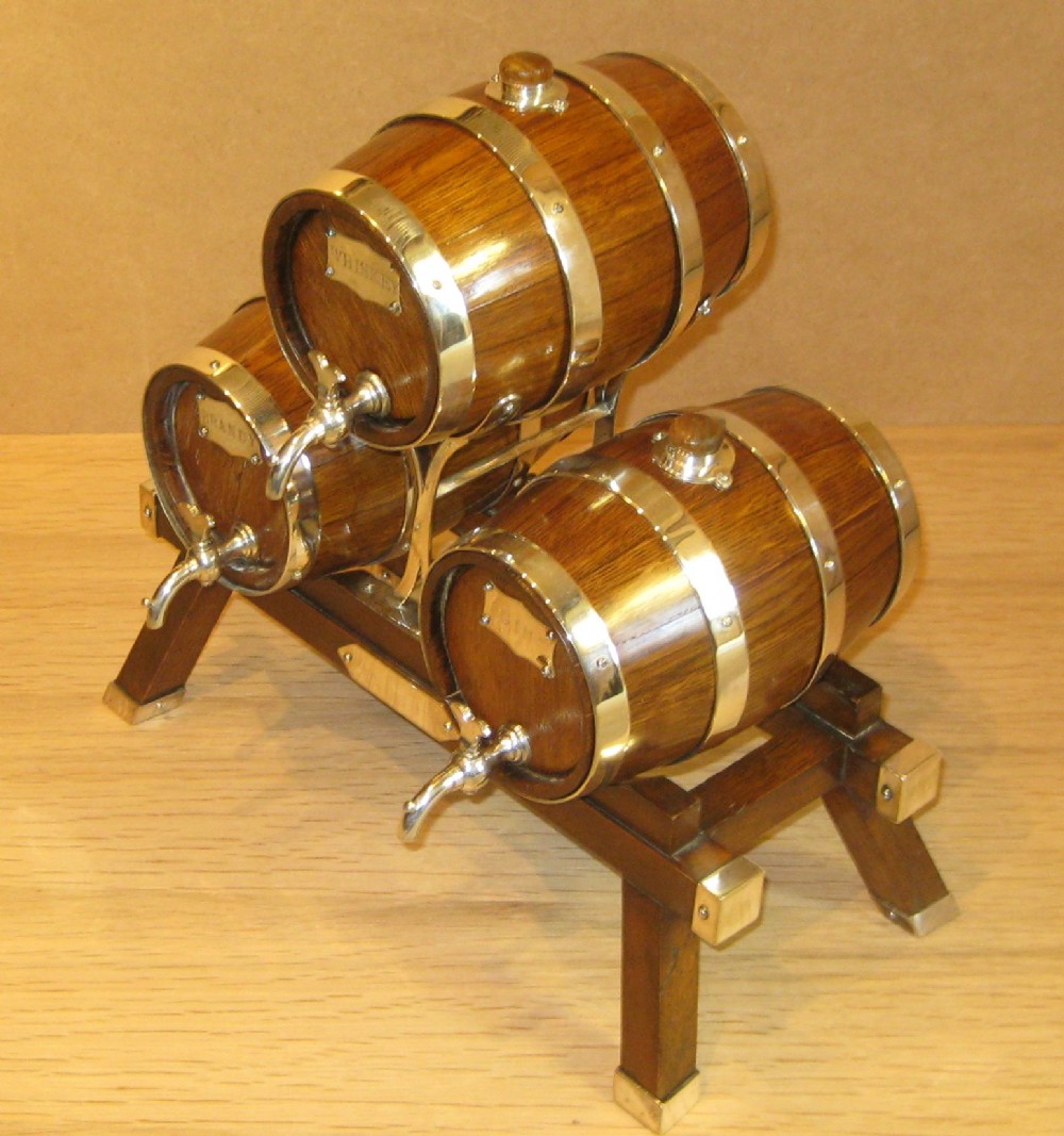 A triple miniature oak spirit barrel dispenser set mounted on a wooden frame with silver-plated hoops, spigots, and metal spirit labels.