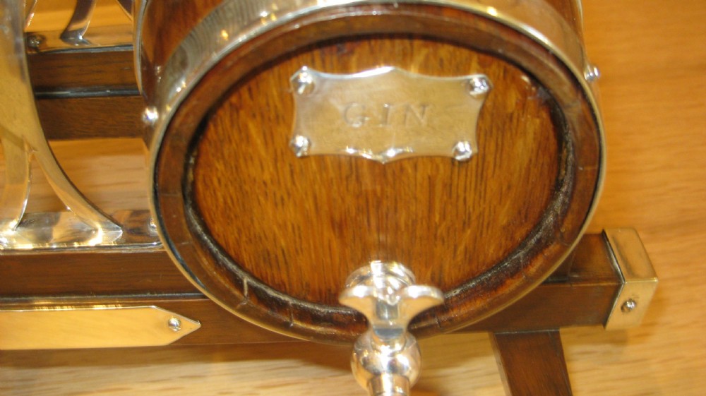 A triple miniature oak spirit barrel dispenser set mounted on a wooden frame with silver-plated hoops, spigots, and metal spirit labels. View V