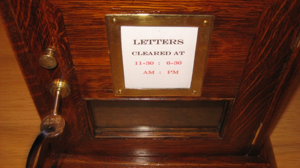 A Victorian oak table-top letterbox featuring an engraved brass top plate with red VR lettering, a glass door panel, and a key inserted in the lock. View III