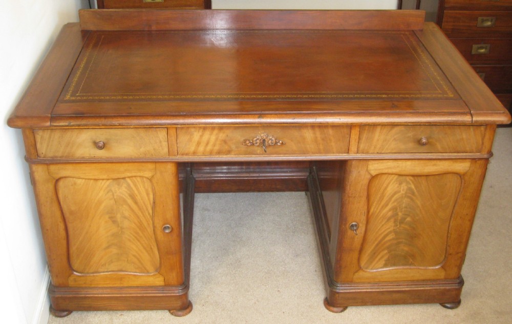 A Victorian faded mahogany pedestal desk featuring a gilt-bordered leather writing surface, three frieze drawers, and twin pedestal cupboards with arched flame mahogany panels. View IV
