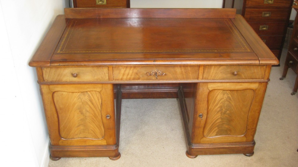 A Victorian faded mahogany pedestal desk featuring a gilt-bordered leather writing surface, three frieze drawers, and twin pedestal cupboards with arched flame mahogany panels.