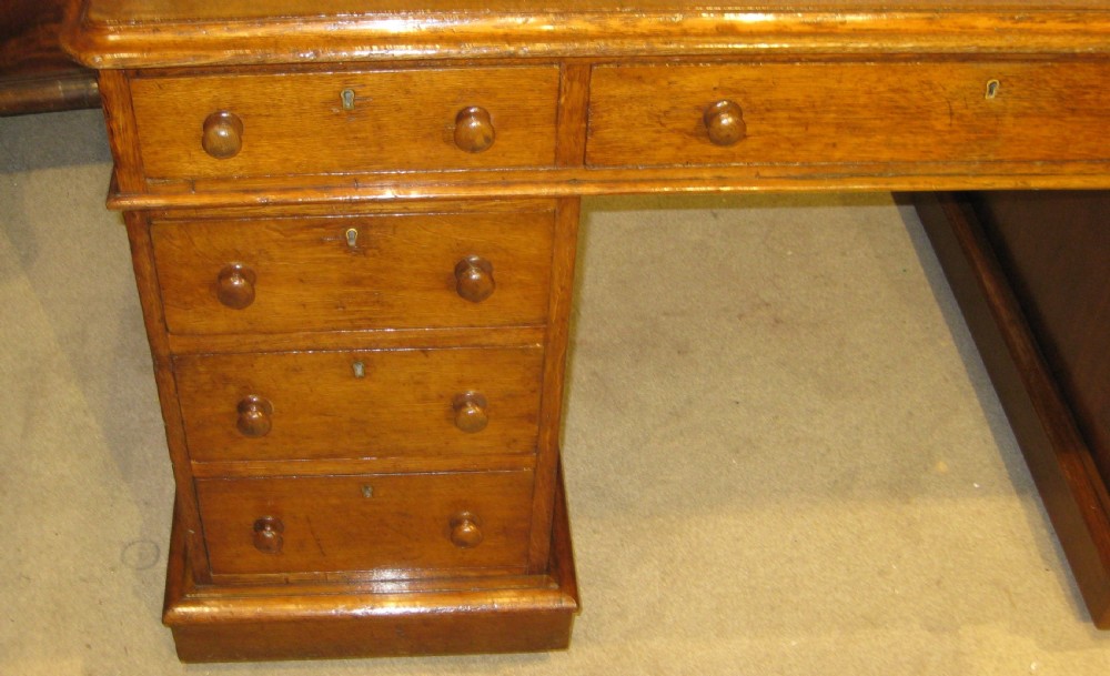 A Victorian golden oak pedestal desk featuring a green leather writing surface, nine drawers with round wooden handles, and plinth bases. View III