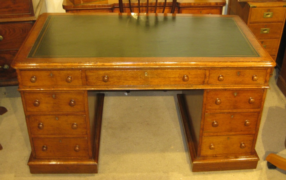 A Victorian golden oak pedestal desk featuring a green leather writing surface, nine drawers with round wooden handles, and plinth bases. View V