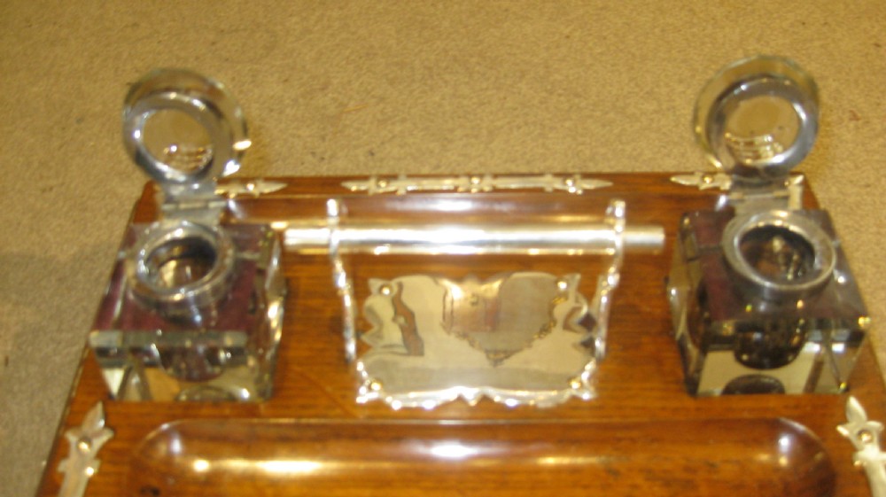 A rectangular Victorian oak inkstand featuring two square glass inkwells, a carved central pen trough, and decorative silver-plated metal strapwork mounts. View V