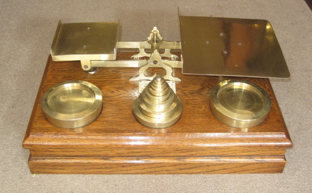An Edwardian postal scale set featuring polished brass pans and stacked brass weights mounted on a moulded solid oak base.