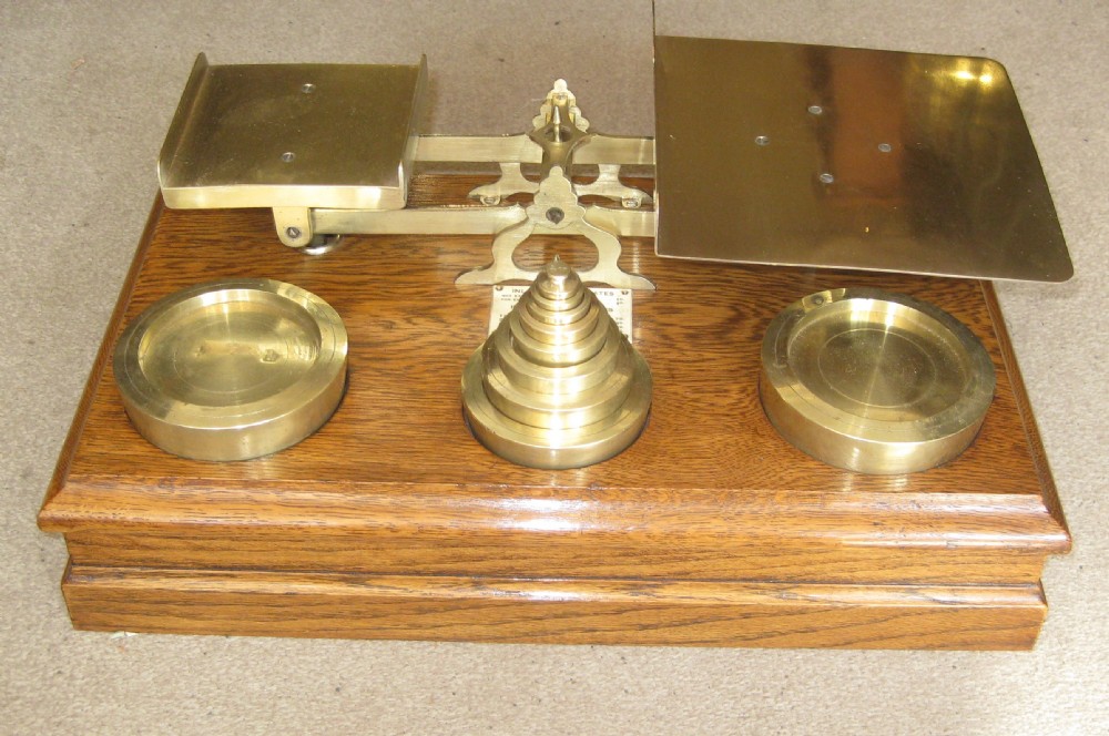 An Edwardian postal scale set featuring polished brass pans and stacked brass weights mounted on a moulded solid oak base. View VII
