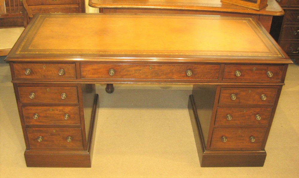 A Victorian mahogany pedestal desk featuring a tan gold-tooled leather top, nine drawers with round brass handles, and plinth bases.