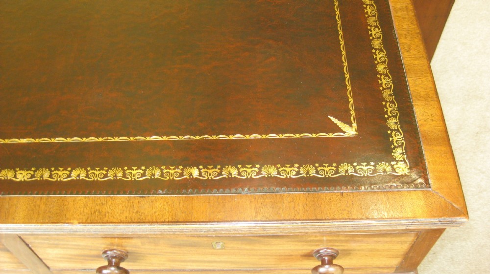 A Victorian mahogany pedestal desk featuring a dark leather writing surface with gold tooling, turned wooden knob handles, and nine storage drawers resting on twin plinth bases. View V
