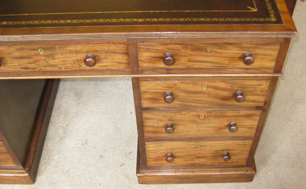 A Victorian mahogany pedestal desk featuring a dark leather writing surface with gold tooling, turned wooden knob handles, and nine storage drawers resting on twin plinth bases. View VII