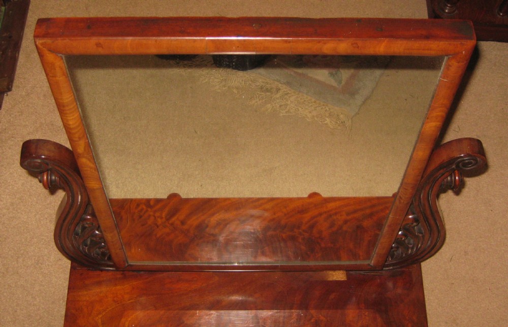 A Victorian flame mahogany swing dressing mirror featuring carved scroll upright supports and a rectangular tilting glass resting on a base with drawers. View II