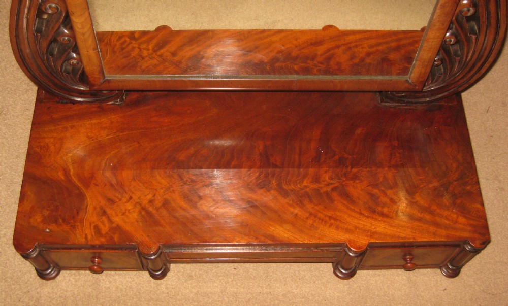 A Victorian flame mahogany swing dressing mirror featuring carved scroll upright supports and a rectangular tilting glass resting on a base with drawers. View IV