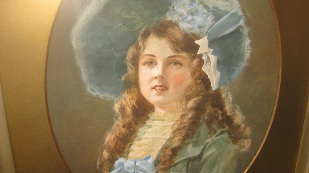 A late Victorian watercolour portrait depicting a young woman wearing a blue hat and dress set within an oval mount and heavily carved gilt frame. View II