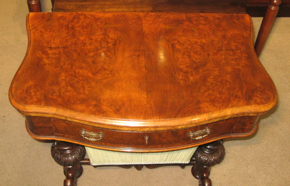 A Victorian burr walnut games and sewing table featuring a serpentine fold-over top, carved turned supports, a single drawer, and a fabric-covered work basket below. View II