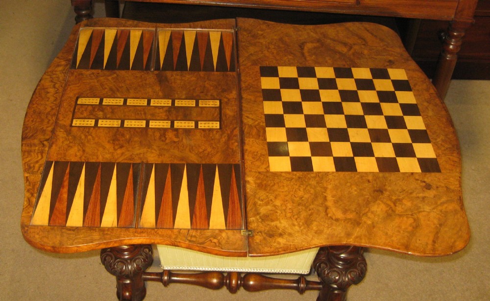 A Victorian burr walnut games and sewing table featuring a serpentine fold-over top, carved turned supports, a single drawer, and a fabric-covered work basket below. View IV