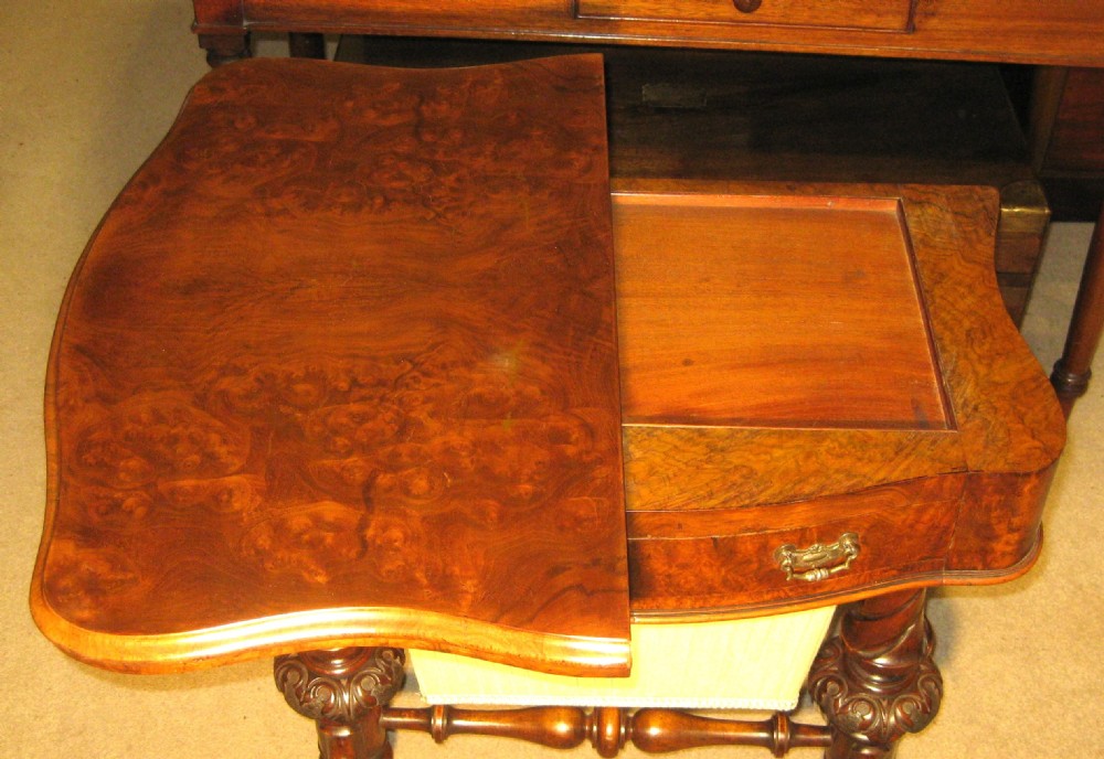 A Victorian burr walnut games and sewing table featuring a serpentine fold-over top, carved turned supports, a single drawer, and a fabric-covered work basket below. View VI