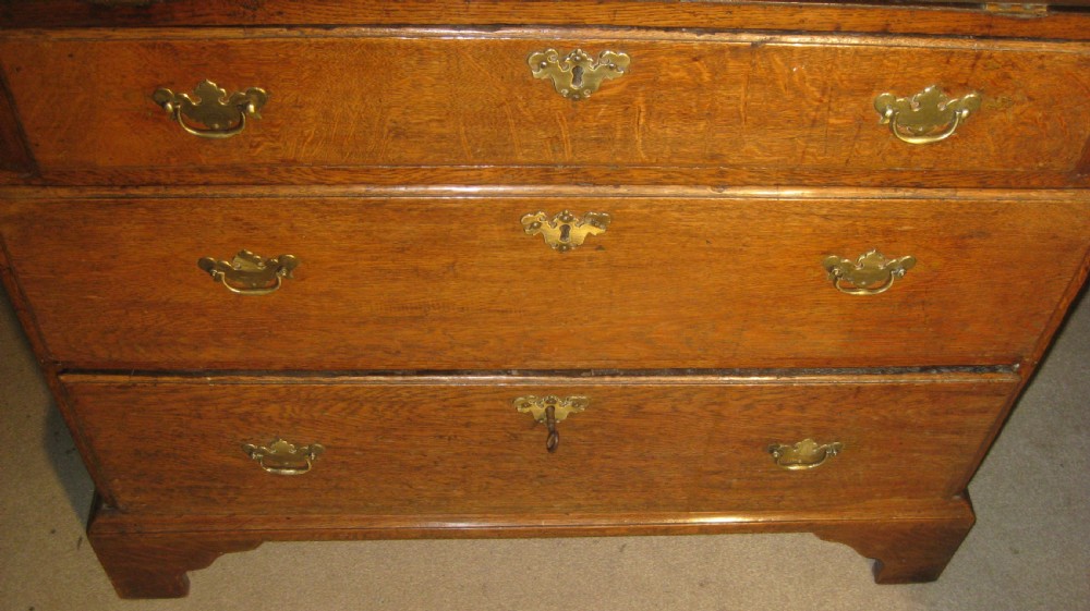 An early Georgian solid oak slant-front bureau featuring three full-width drawers with brass swing handles and bracket feet. View II