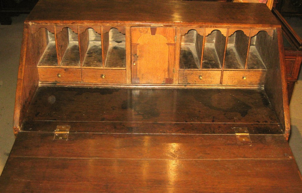 An early Georgian solid oak slant-front bureau featuring three full-width drawers with brass swing handles and bracket feet. View III
