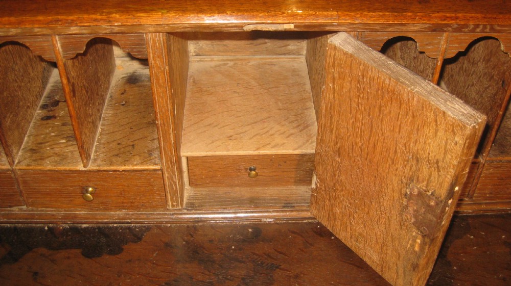 An early Georgian solid oak slant-front bureau featuring three full-width drawers with brass swing handles and bracket feet. View V