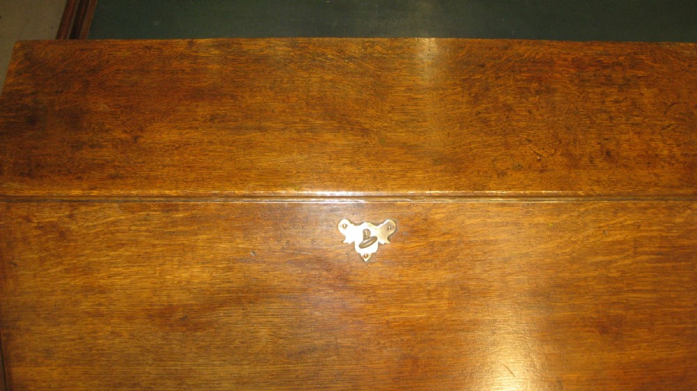 An early Georgian solid oak slant-front bureau featuring three full-width drawers with brass swing handles and bracket feet. View VII