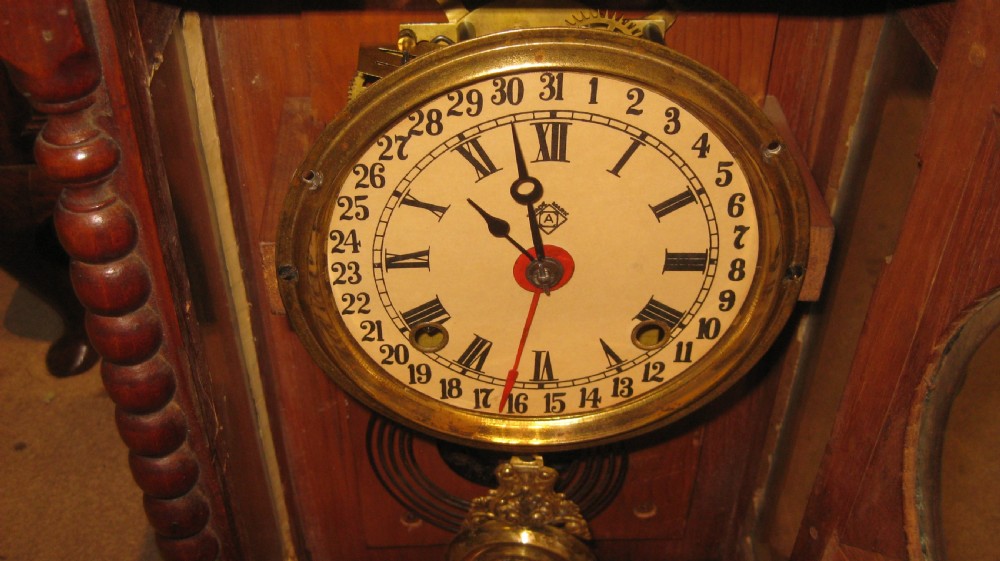 An Edwardian walnut mantel clock featuring bobbin-turned side columns, a cream calendar dial, and a glass lower panel showing a brass pendulum. View II