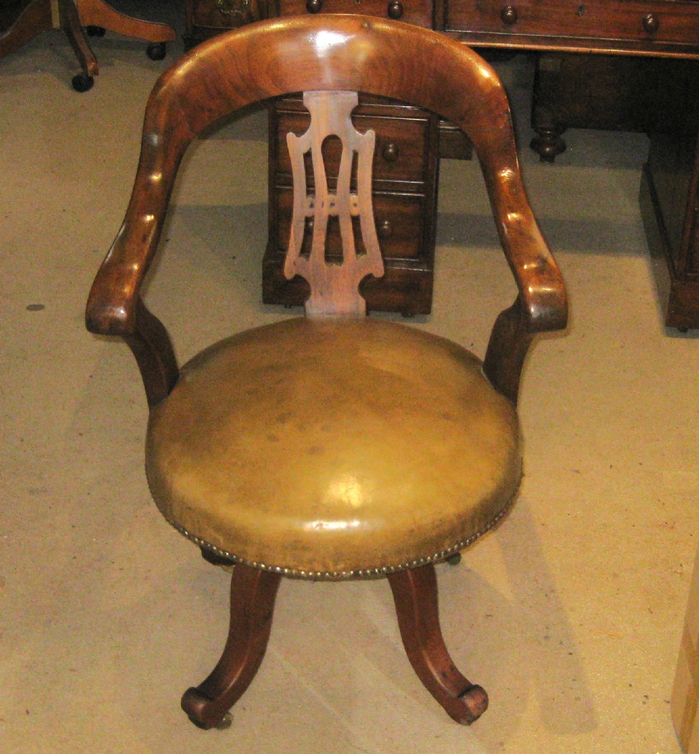A Victorian walnut revolving desk chair featuring a curved horseshoe backrest, pierced carved splat, circular tan leather upholstered seat with brass studding, and four splayed legs on castors.