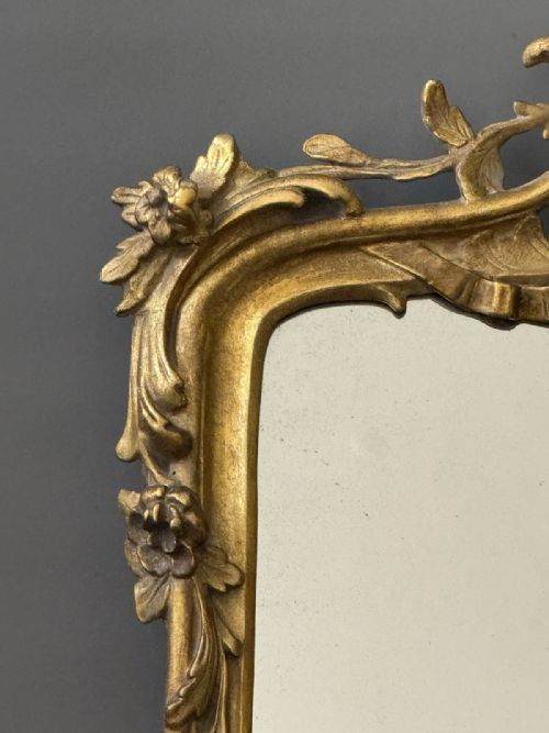 fig 1. A carved French Rococo giltwood wall mirror featuring a detailed scrolling crest, floral side accents, and a rectangular glass plate.