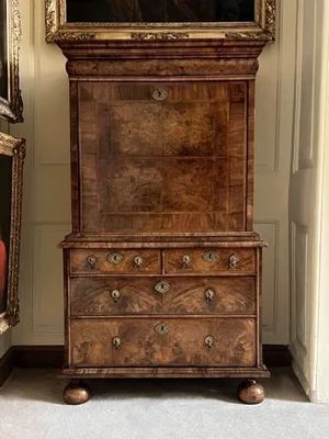 fig 2. An antique William and Mary walnut secretaire chest featuring figured walnut veneers, a fall-front upper section, multiple drawers, and turned bun feet.