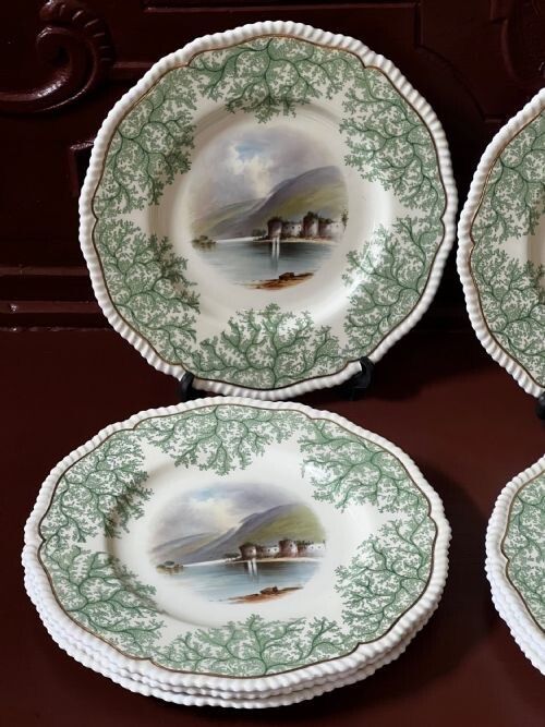 fig 1. A set of English ceramic dinner plates featuring green seaweed borders, scalloped gilt rims, and central hand-painted landscape scenes depicting castles near water.