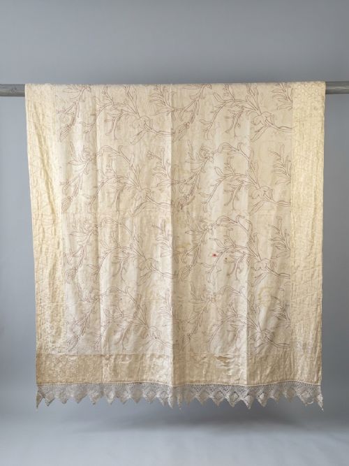fig 1. A cream-coloured Art Nouveau silk coverlet featuring hand-embroidered botanical motifs with a quilted border and pointed lace trim.