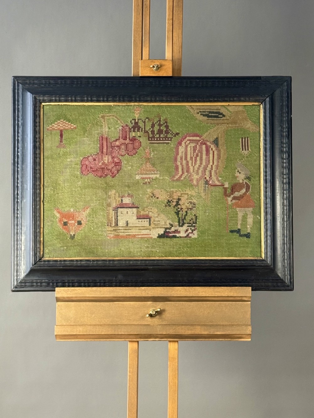 A framed antique needlework panel featuring an olive green wool ground stitched with naive figures, a fox head, trees, and buildings inside a dark ebonised wooden frame.