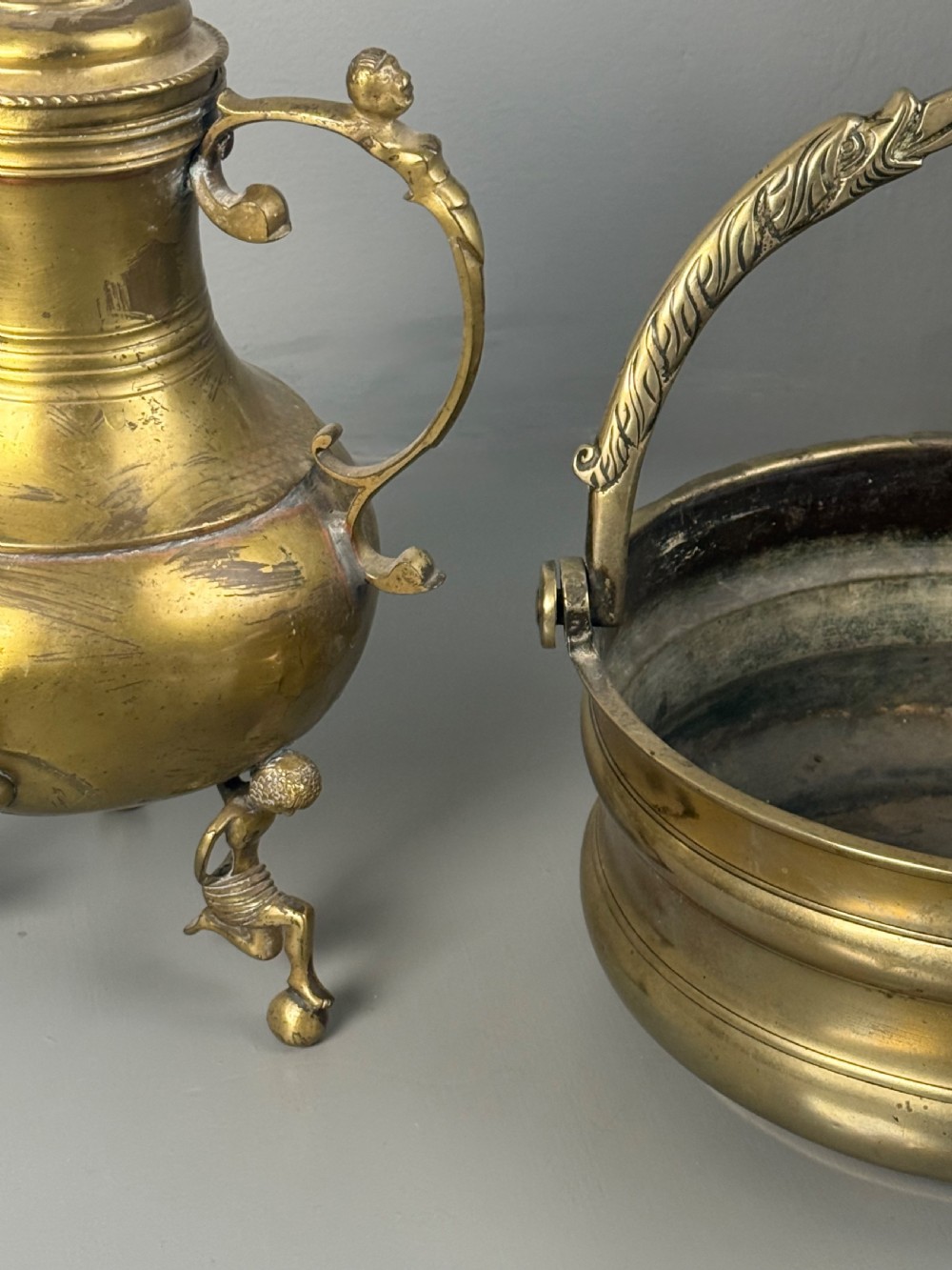 An aged brass urn with caryatid supports and a top finial lid, displayed alongside a brass vessel featuring a raised dragon-form handle. View VI
