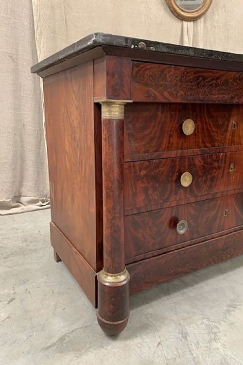 fig 1. A nineteenth-century French Empire flame mahogany commode featuring three drawers, side column supports, circular brass handles, and a dark marble top.