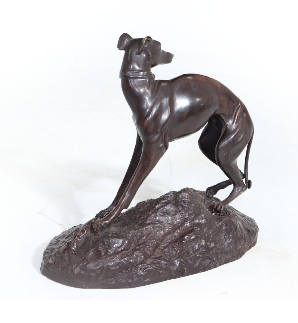 A dark brown patinated bronze sculpture of a standing whippet dog on an uneven textured base.