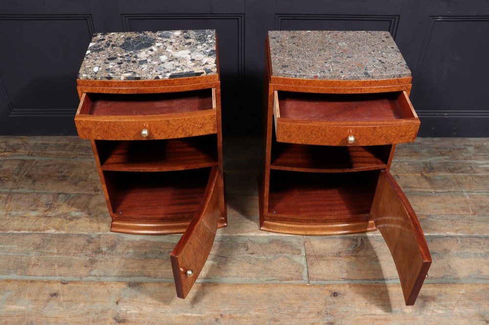 A pair of Art Deco amboyna wood bedside cabinets featuring patterned stone tops, single drawers, open shelf compartments, and curved cupboard doors.