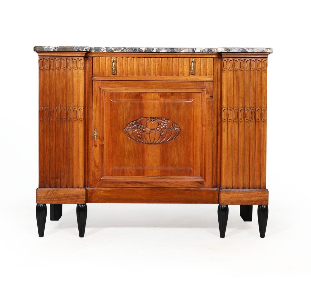 A French Art Deco walnut sideboard featuring a grey mottled marble top, fluted side panels, a central carved fruit motif door, and black turned feet.
