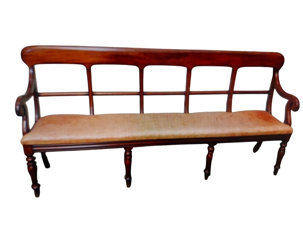 A long nineteenth-century mahogany bench seat featuring turned front legs, scrolled armrests, a multi-panel open backrest, and a tan upholstered seat.
