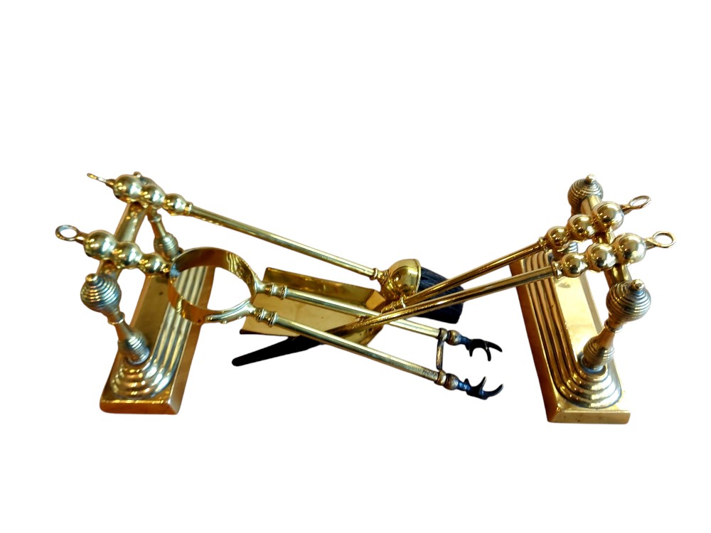 A small polished brass fireside companion set featuring two firedogs with turned finials and resting hand tools.