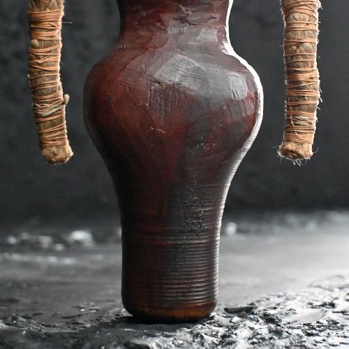 fig 1. A carved wooden folk art figure featuring facial details, wrapped textile arms, and a tapered cylindrical base.