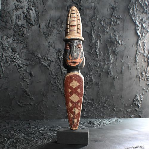 fig 1. A carved wooden whirligig figure featuring a stylized human head with a ribbed hat and geometric painted patterns along the lower torso.