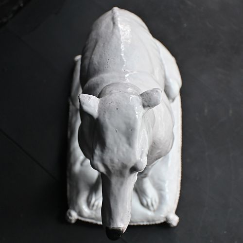 fig 1. A white glazed ceramic statue depicting a slender, seated greyhound dog resting on a tasselled cushion pedestal.