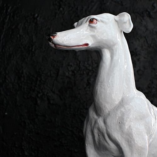 fig 3. A white glazed ceramic statue depicting a slender, seated greyhound dog resting on a tasselled cushion pedestal.