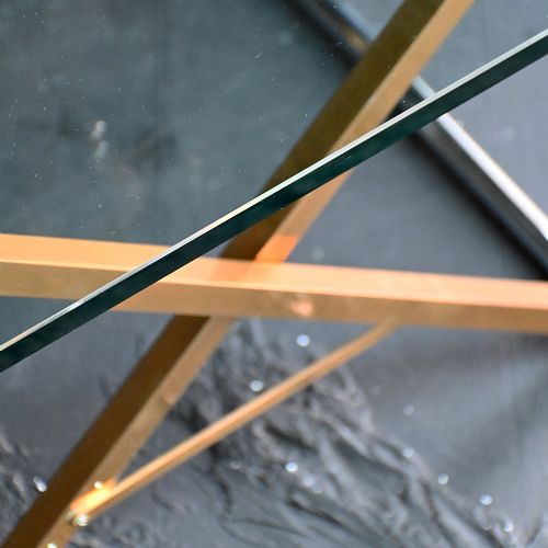 fig 2. A square clear glass side table supported by a brass and metal X-frame base.