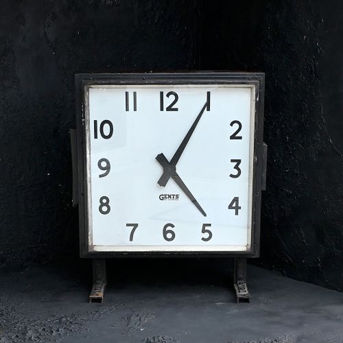fig 1. A square black steel Gents of Leicester station clock featuring a white face with bold black Arabic numerals and metal mounting feet.