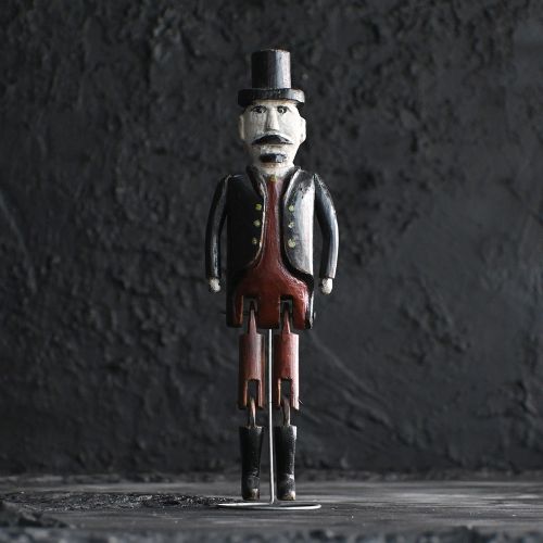 fig 1. A carved wooden jigger doll with jointed legs, wearing a black coat, top hat, brown waistcoat, and boots against a plain background.