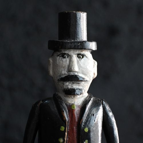 fig 2. A carved wooden jigger doll with jointed legs, wearing a black coat, top hat, brown waistcoat, and boots against a plain background.