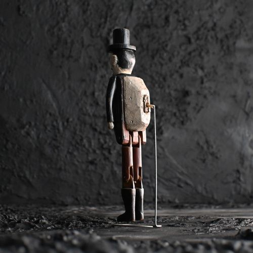 fig 3. A carved wooden jigger doll with jointed legs, wearing a black coat, top hat, brown waistcoat, and boots against a plain background.